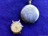 Front view of the antique Dueber Coin silver pocket watch beside the gold-toned wristwatch showing dial and ornate case engraving