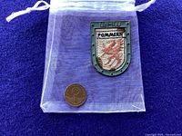 Photo of Pommern Tinnie badge and vintage coin placed inside a small translucent plastic bag on blue fabric background.