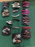Top down photo of 15 assorted Karina vintage hair clips and barrettes arranged on a green cloth, still mounted mostly on black Karina brand cards with some loose items.