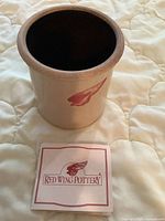 Stoneware crock with Red Wing logo displayed on a quilted surface, with informational inserts about Red Wing Pottery placed in front.