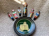 Seven hand painted lacquered wooden dolls and one decorative wooden plate with Cyrillic writing, arranged in a fan shape on a textured surface.