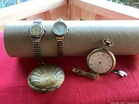 Three watches and two tie clips displayed on red cloth, pocket watch gold-tone with engraved cover, two wristwatches with metal bands.