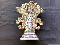 Ornate ceramic vase from Coimbra with multi-color floral and ornamental hand painted designs on cream background, scalloped top and curved sides, placed on black fabric surface.