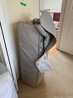 View of the folded portable bed in a grey fabric zipped carry bag, standing upright against a wall.