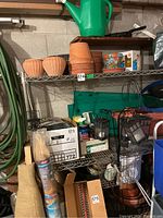 Wide view of metal shelf with watering can on top, terracotta pots, decorative painted flower pots, bird netting, outdoor candle lanterns, gardening accessories and yard bag support.