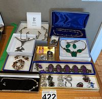 Full lot layout showing various costume jewelry pieces arranged in boxes and trays including Bastien Joaillier set, green stone set, amethyst bracelet, pearl and pink necklaces, and assorted earrings and rings.