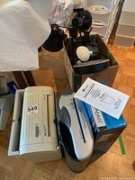 Multiple paper shredders of different colors and styles, with a beige model accompanied by paperwork/manuals, shown alongside the box of lamps.