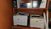 Two HP LaserJet printers on shelf under shelf with ink cartridges