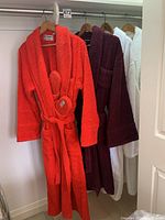 Four terrycloth robes hanging on wooden hangers in a closet, including one bright red robe with matching slippers.