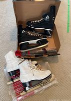Two pairs of ice skates in a cardboard box with protective packaging. One pair is black Daoust National men's skates, the other is white women's figure skates.