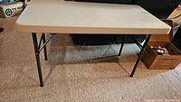 Full view of the beige rectangular folding table with black metal legs set up indoors on carpet.