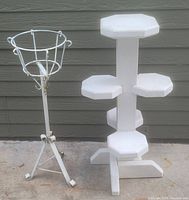 Two white plant stands side by side against an outdoor wall. One metal with basket top and tripod base; the other wooden with multiple octagonal shelves.