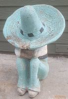 Front view of the concrete figurine showing the large turquoise wide-brimmed hat, with arms resting on knees.