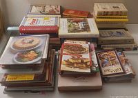 Top-down view showing multiple stacks of cookbooks with different covers and sizes, illustrating variety and quantity.
