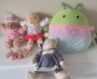 Four dolls and plushies displayed against a wall including two Cabbage Patch Kids dolls, a pastel round plush bug, and a teddy bear in dress.