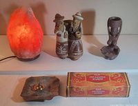 Photo showing Himalayan salt rock lamp glowing, two ceramic figurine incense holders shaped like a man and woman in traditional attire, a wooden figurine incense holder shaped like a figure, and a box of Dragon's Blood incense sticks with a brass holder dish.