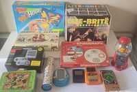 Photo showing various vintage kids games and toys including boxed Lite-Brite sets, Spirograph, Nerf Pro Hoop basketball game, Tamagotchi device, and several handheld electronic games.