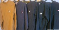 Six men's long sleeve shirts and sweatshirts on hangers against a wall, from front view showing logos
