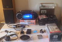 Wide view of all electronics and accessories including speakers, boom box, cassette player, VHS and DVD players, headphones, microphone, USB drive, and boxed media rack.
