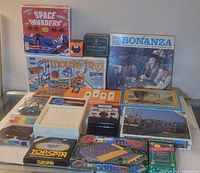 Photo showing various boxed board games and puzzles arranged on a shelf including Space Invaders, Bonanza, Mouse Trap, Top Spin, and several puzzles