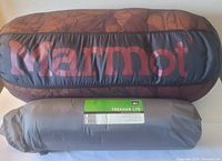 Rolled Marmot sleeping bag in printed stuff sack alongside rolled REI pad in grey sack.