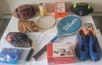 Photo of entire sports lot showing two footballs, two baseball gloves, tennis racquet, blue athletic shoes, goggles, can of tennis balls, timer clock, and box of foot care product