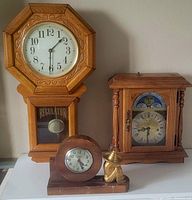 Frontal view of three vintage clocks on table: octagonal regulator wall clock, wooden mantel clock with figurine, rectangular mantel clock with moon phase dial.