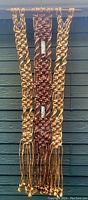 Full view of vintage rope macrame wall hanging with three vertical strands in brown tones, hung against a wooden wall background