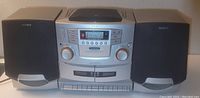 Sony portable stereo boom box showing front view with two black speakers attached on sides. Unit has large volume and tuning knobs, cassette and CD compartments, and digital display showing radio station.