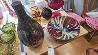 Photo of the full lot showing a large mosaic glass vase and a colorful ceramic plate on a wooden table alongside other glass items.