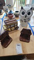 Multiple ceramics on a table including the owl cookie jar, wagon cookie jar, blue and white vases and cups, and two brown bookends.