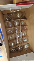 Box containing 14 handblown glass Christmas ornaments divided in cardboard compartments showing a variety of spherical and icicle shaped ornaments with gold and iridescent finishes.