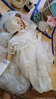 Full body photo of doll lying on a surface, dressed in a white lace baptismal gown with bonnet and sheer veil.