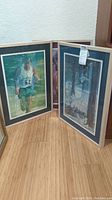 Three framed sports-themed art prints leaning against a wall on a wooden floor. The art depicts athletes, one clearly a runner with bib number 62. All pieces have mats and glass frames.