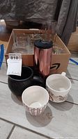 Lot showing coffee mugs and a copper colored insulated tumbler in front of a box containing clear glass tumblers.