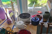 Kitchenware collection displayed on wooden table with natural light from window. Includes white ceramic teapot, glass pump bottle, purple and amber glass vessels.