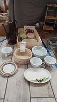 Full set of kitchenware including bowls, cups, plates, multi-colored plate, pedestal bowl, and dark metal chocolate pot arranged on wooden floor.