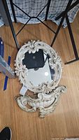 Oval ornate mirror and matching decorative piece on wooden floor, showing elaborate white and gold carved frames.