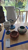 Photo of four pottery items on table by window, including tall pitcher, goblet, two vases.