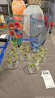 Photo shows five small drinking glasses decorated with yellow flowers and two taller glass vases. One vase is clear with multi-colored flowers painted on it, the other is frosted white.