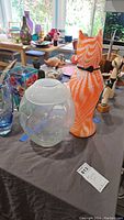 Two vases on table: clear Waterford crystal vase with rounded shape and flared rim, next to orange and white swirled cat-shaped art glass vase with black collar.