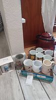 Box of assorted coffee cups including floral porcelain cups and novelty mugs arranged on the floor with metal tea pot in the background.