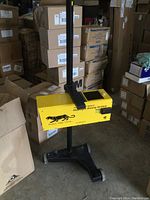 Full front-angle view of yellow headlight aiming machine on black stand surrounded by warehouse boxes