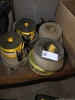 Eight wide tow straps rolled up, including two bright yellow Kennedy brand straps with metal hooks and six worn beige straps, arranged on a wooden surface.