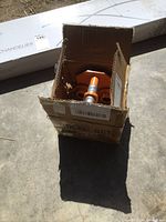 Open cardboard box containing orange metal hand trolley parts with threaded shafts and wheel assembly.