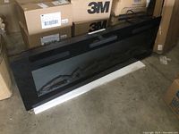 Front view of black electric wall mount fireplace heater showing ceramic logs inside glass panel, resting on white foam base against cardboard boxes.