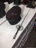 Side view of fencing mask showing black metal mesh and attached white neck protector with fencing foil placed next to it.