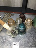 Image showing six antique insulators on metal surface, including one with attached wooden post, glass and ceramic materials, varying colors.