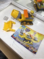 Yellow and gray Mega Bloks cement mixer vehicle with Chase figure, yellow scoop attachment, white blocks and instruction booklet visible on white surface