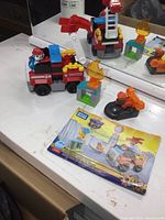 Photo showing the assembled Paw Patrol fire truck with firefighter figure inside, accessories including two flame pieces and small orange rescue vehicle, and the colorful instruction booklet on white surface in front of mirror.